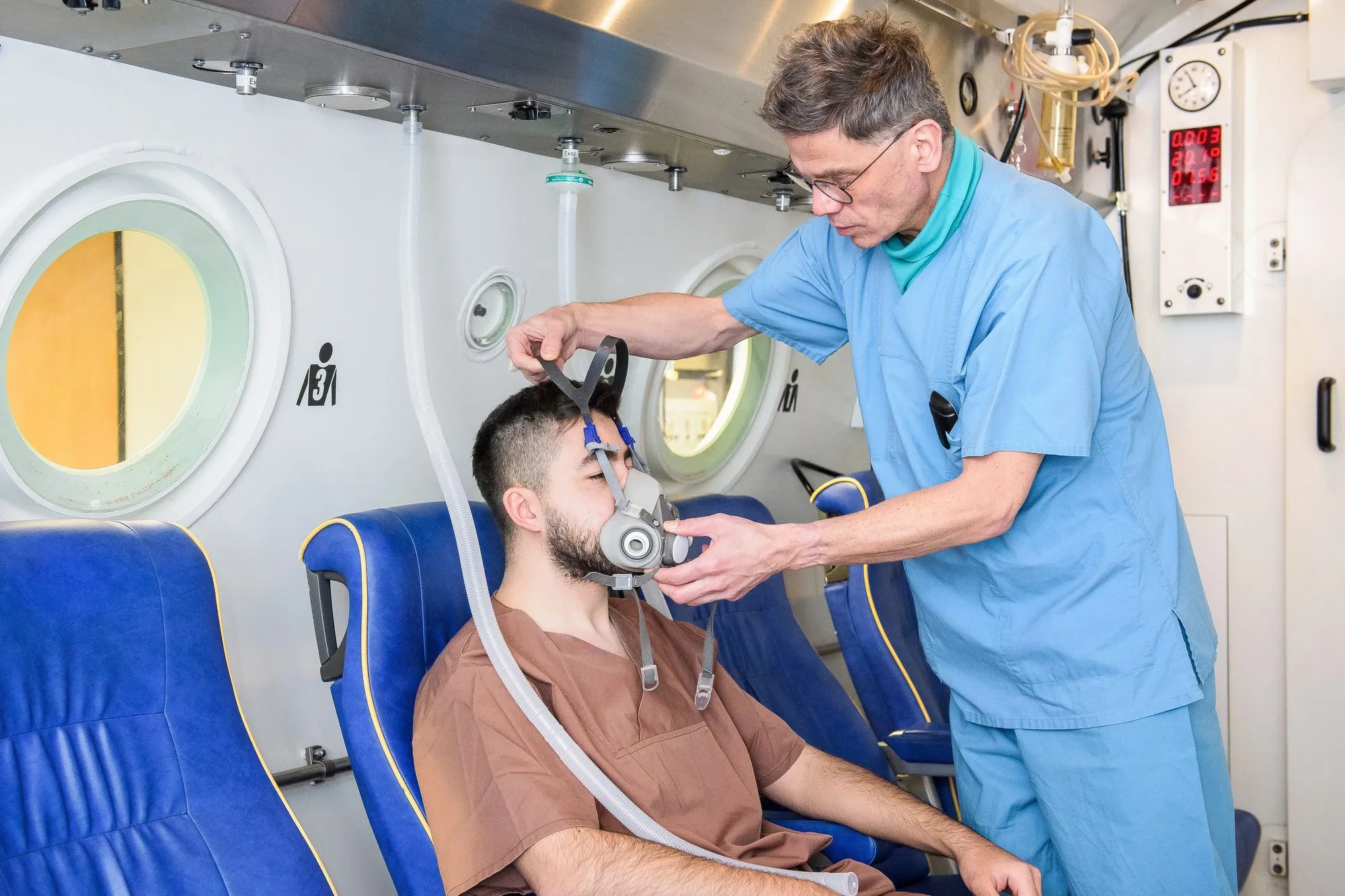 Ein Mitarbeiter legt einer Person in der Mehrplatz-Druckkammer die Atemmaske an, Zentrum für Hyperbarmedizin am Universitätsklinikum Halle (Saale)