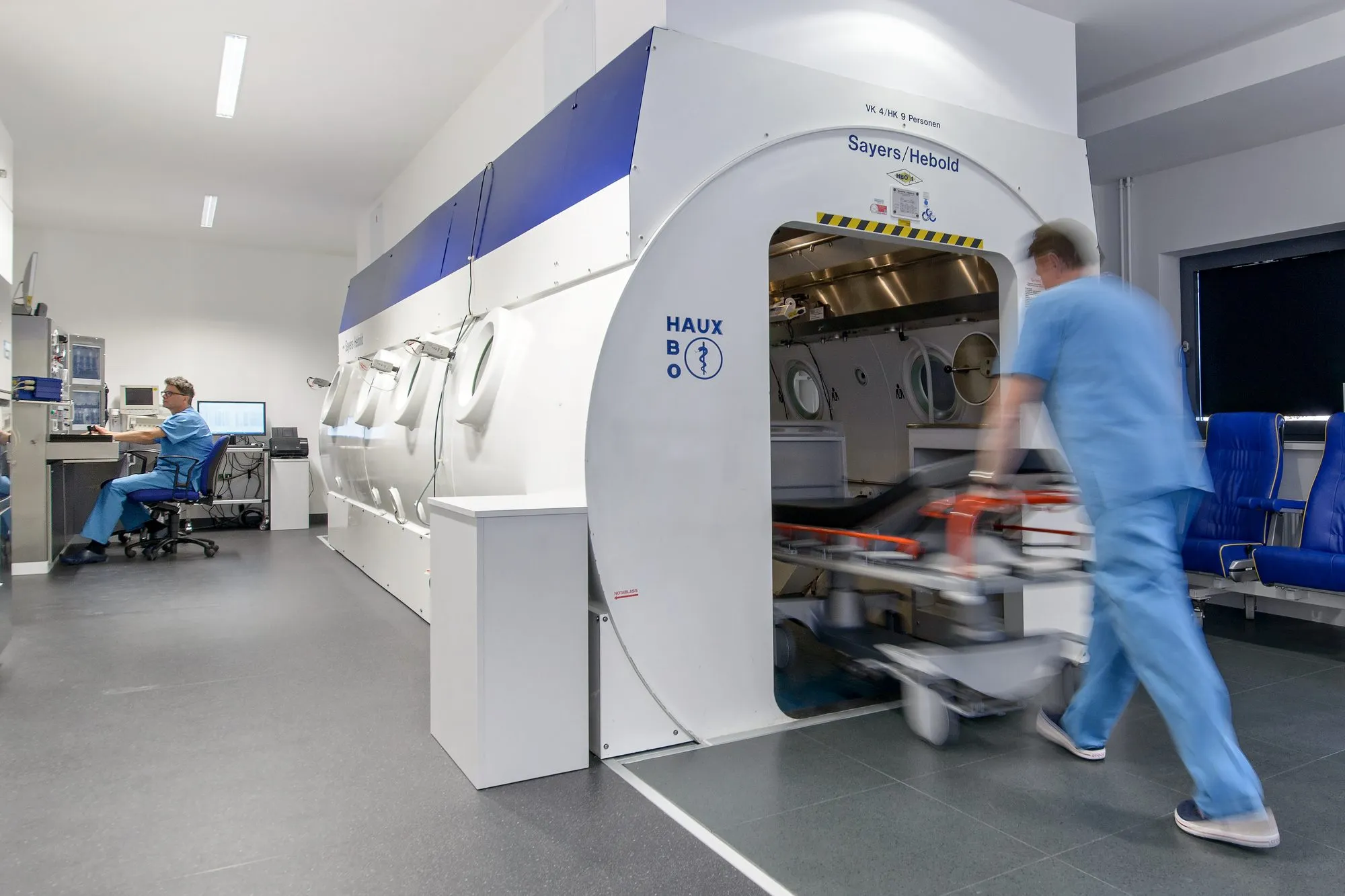 Behandlungsraum mit Mehrplatz-Druckkammer und Leitstand; ein Mitarbeiter sitzt am Leitstand, ein zweiter schiebt eine Trage in die Kammer, Zentrum für Hyperbarmedizin am Universitätsklinikum Halle (Saale)